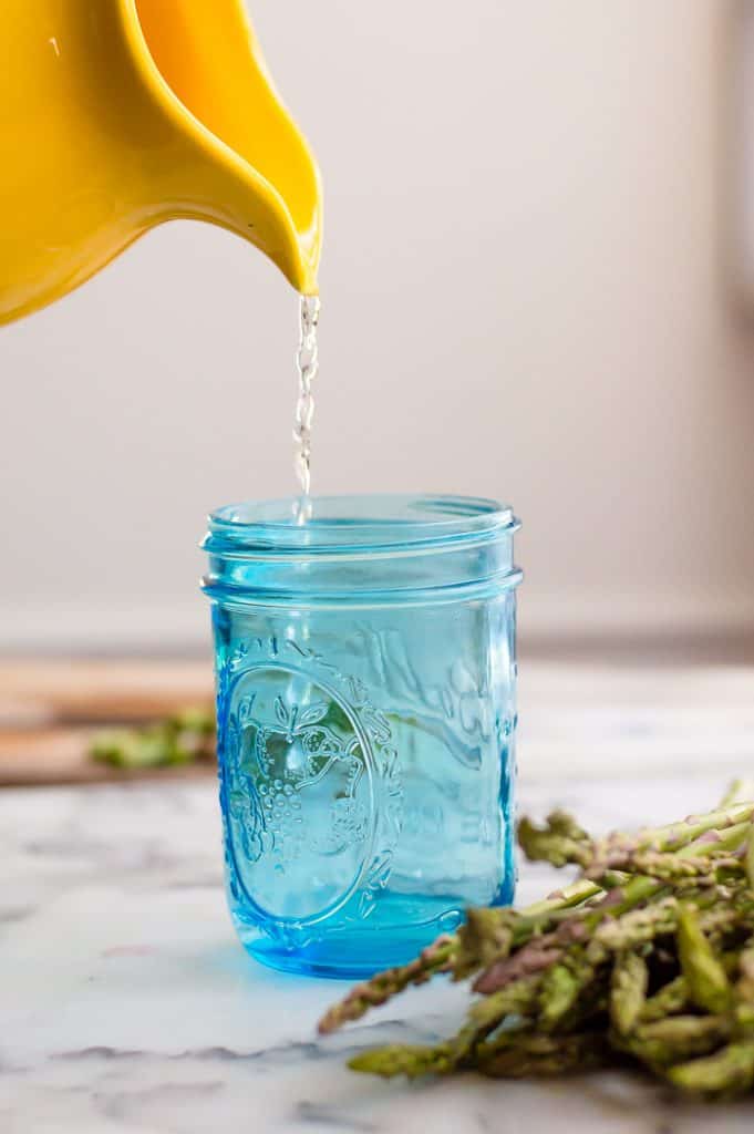 A yellow pitcher pouring water into a blue mason jar. There is fresh asparagus on the table next to the jar.
