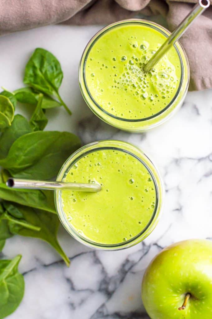 Two glasses of green apple spinach smoothie with metal straws. There is a green apple and spinach leaves on the table next to it the glasses. 