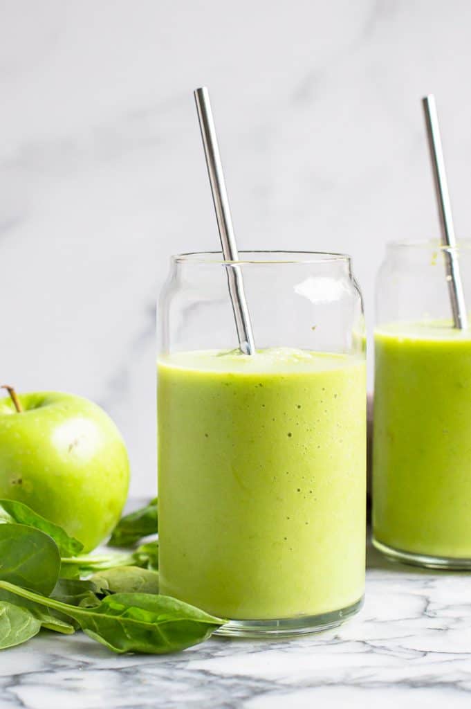 Spinach green apple smoothie in two glasses with metal straws on a table with spinach and a green apple next to them. 