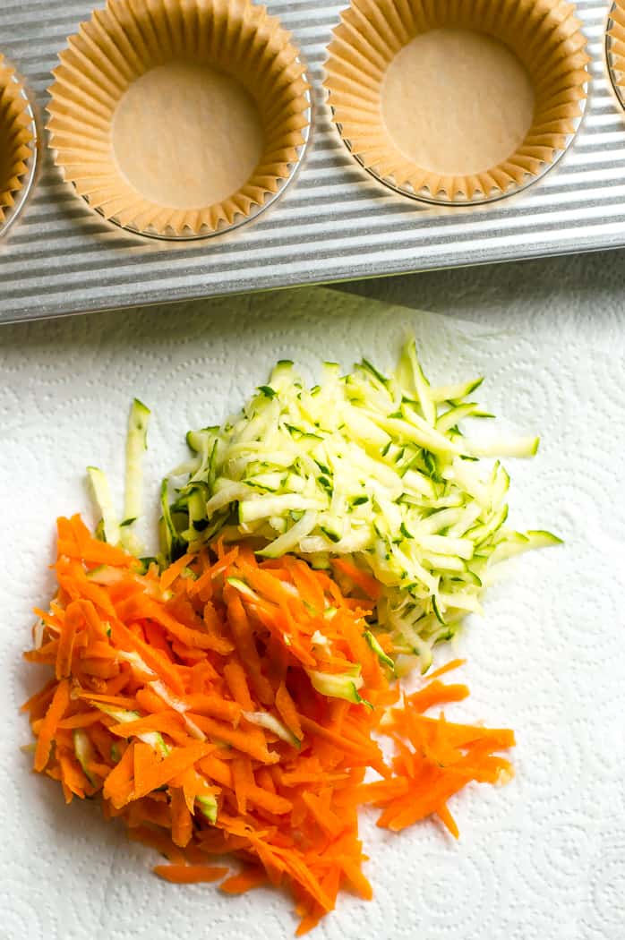 Grated carrots and zucchini in the middle of a paper towel. There is a muffin tin next to the towel. 