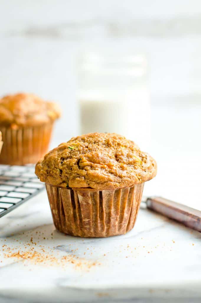 A carrot zucchini muffin on a marble table with a knife next to it and blurred muffins in the background.