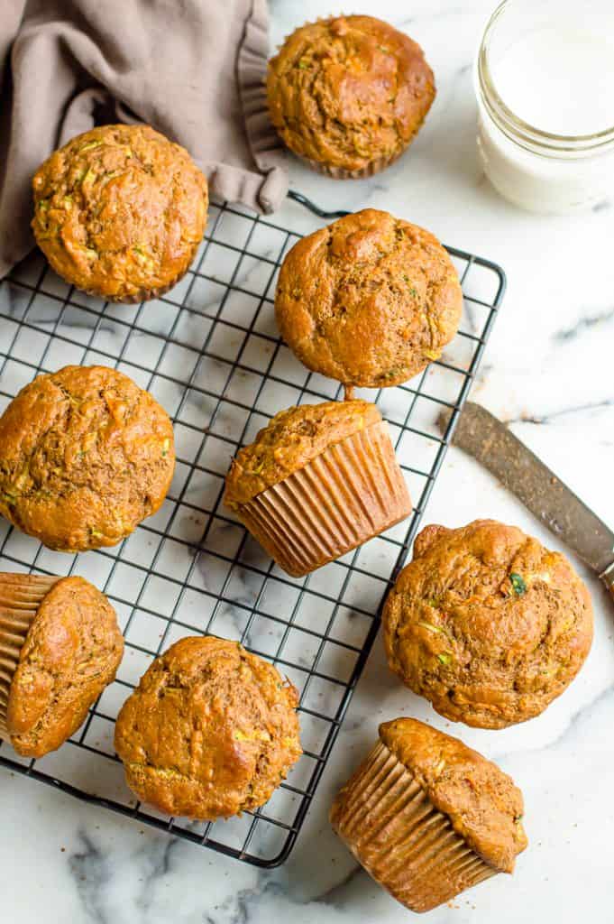 Carrot zucchini muffins on a cooling rack on a white marble table. There is a glass of milk and a butter knife on the table.