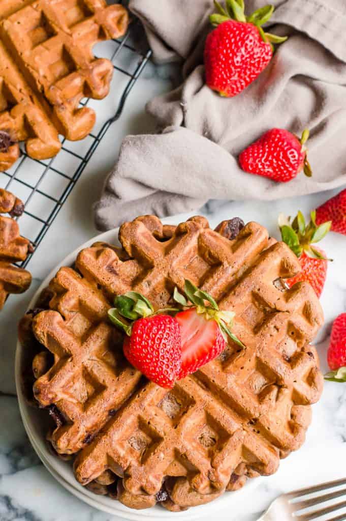 Healthy chocolate waffles on a plate with a sliced strawberry on top. There are more fresh strawberries scattered  around the plate with more waffles next to the plate.
