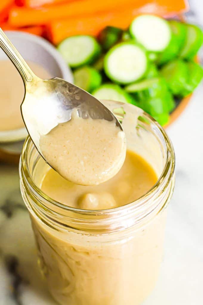 Creamy honey mustard dressing in a jar with a spoon. There are sliced cucumbers and carrots blurred in the background.