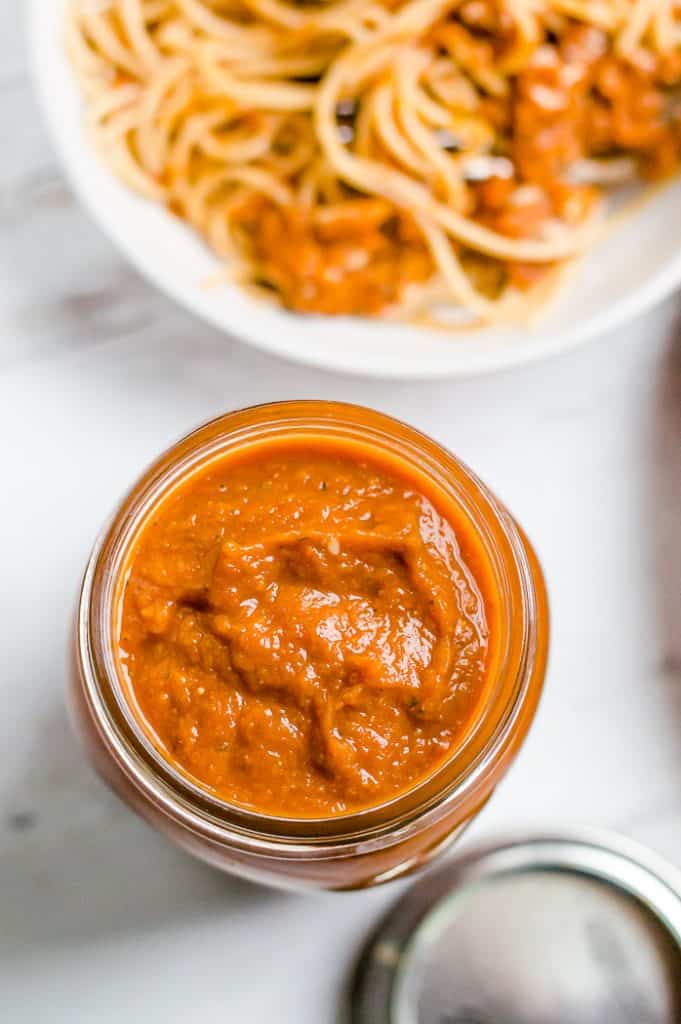 Veggie pasta sauce in a mason jar. There is the jar for the lid next to the jar and a blurred bowl of spaghetti next to it. 