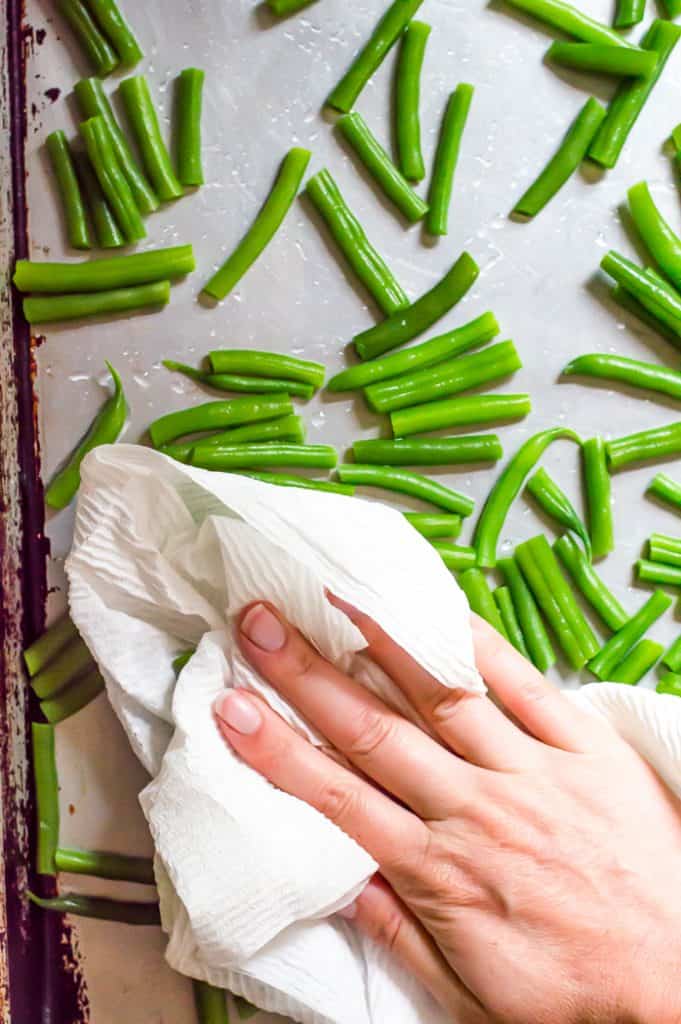 Blanched green beans on a baking sheet with a hand patting them dry with a paper towel. 