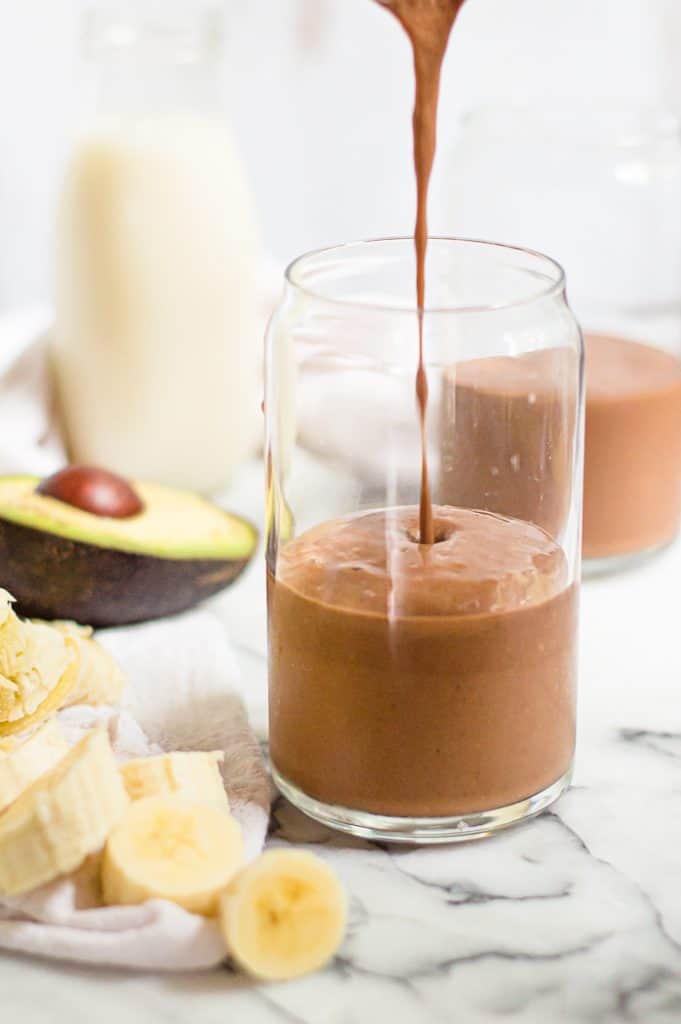 Chocolate avocado banana smoothie being poured into a glass. There is a sliced banana and halved avocado next to the glass and a blurred glass of smoothie in the background. 