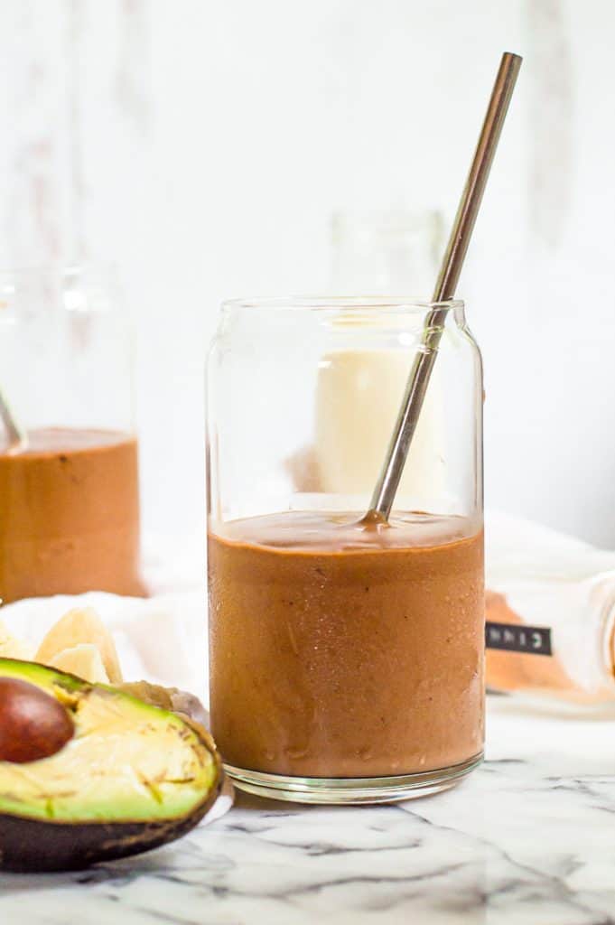 Chocolate avocado banana smoothie in a glass with a metal straw.There is an avocado and a shaker of cinnamon next to eh glass and another glass of smoothie blurred in the background. 