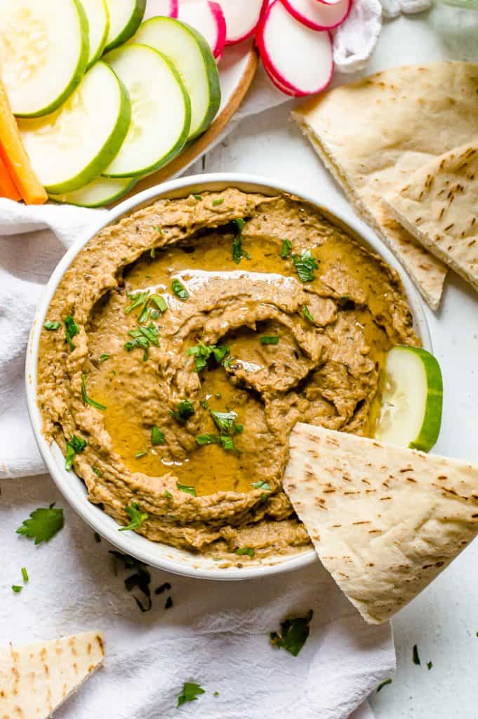 Roasted eggplant dip in a bowl topped with oil and chopped parsley. There is a cut pita and cucumber slice in the bowl with the dip and there are cut veggies and more pieces of pita next to the bowl.
