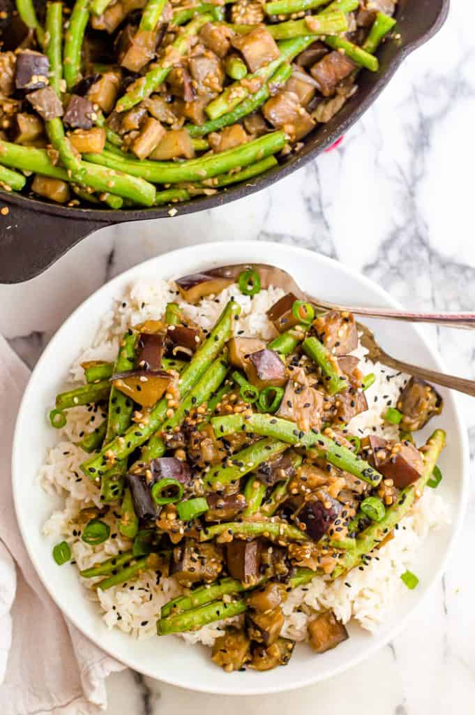 A plate of green bean and eggplant stir fry in a bowl over white rice. The skillet with the rest of the stir fry is next to the plate