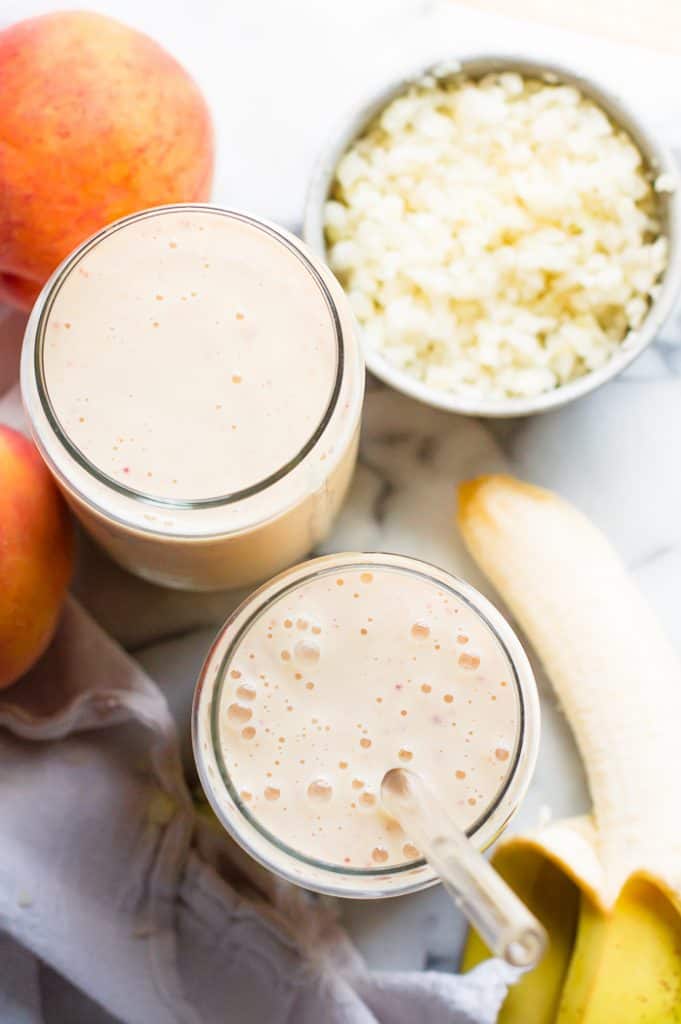 Cauliflower banana peach smoothie in two glasses with peaches and banana next to the glasses. There is a glass straw in one of the glasses.