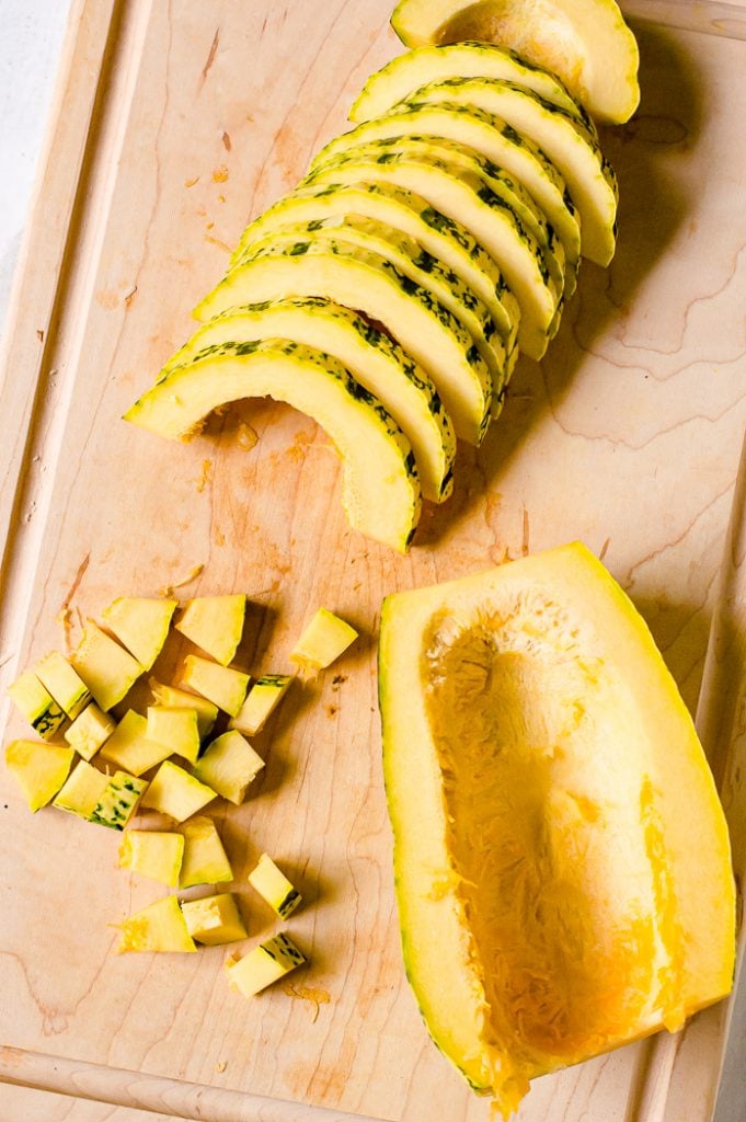 Delicata squash on a cutting board. It has been cut in half and deseeded. One half has been cut into slices and some slices have been diced.