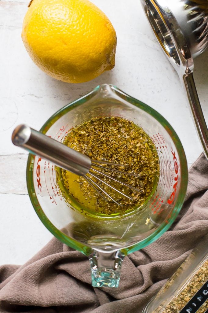 Lemon herb marinade for sheet pan chicken and veggies in a large measuring cup with a whisk. There is a lemon, juicer, grey napkin and jar of dried herbs next to the measuring cup.