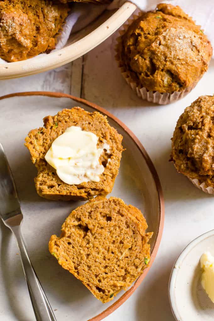 A pumpkin zucchini muffin cut in half on a plate. One half has a little butter on it and there are more muffins on the table next to the plate.