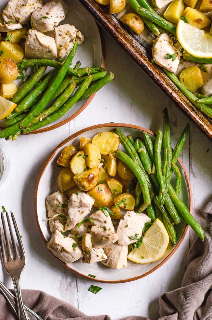 Sheet pan lemon herb chicken green beans and potatoes on two plates on a table with the sheet pan next to it. There is a fork and grey napkin next to the plates.