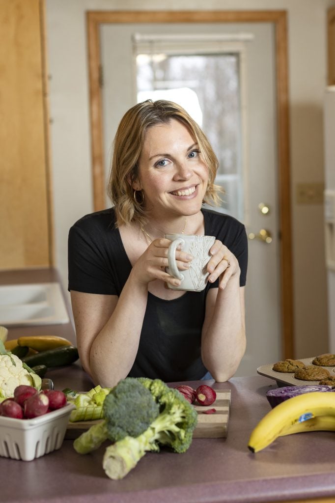 Woman holding a mug with produce and muffins on the counter in front of her. The produce on the counter includes broccoli, bananas, a container of radishes, celery, cabbage and cauliflower.