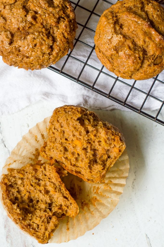 A sweet potato muffin cut in half to show the muffin's texture. The muffin is resting on the liner and is next to a cooling rack with more sweet potato muffins.