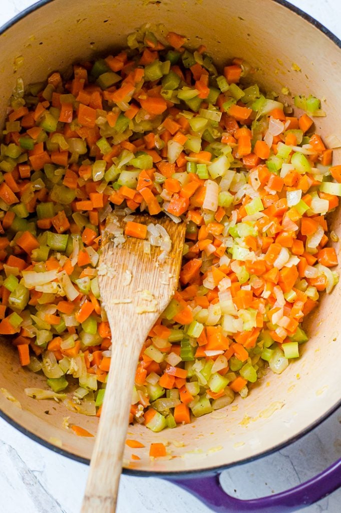 Blue enamel pot on white marble counter with diced carrots, celery, and onion. Wooden spatural is in the pot with bits of the vegetables on it. 