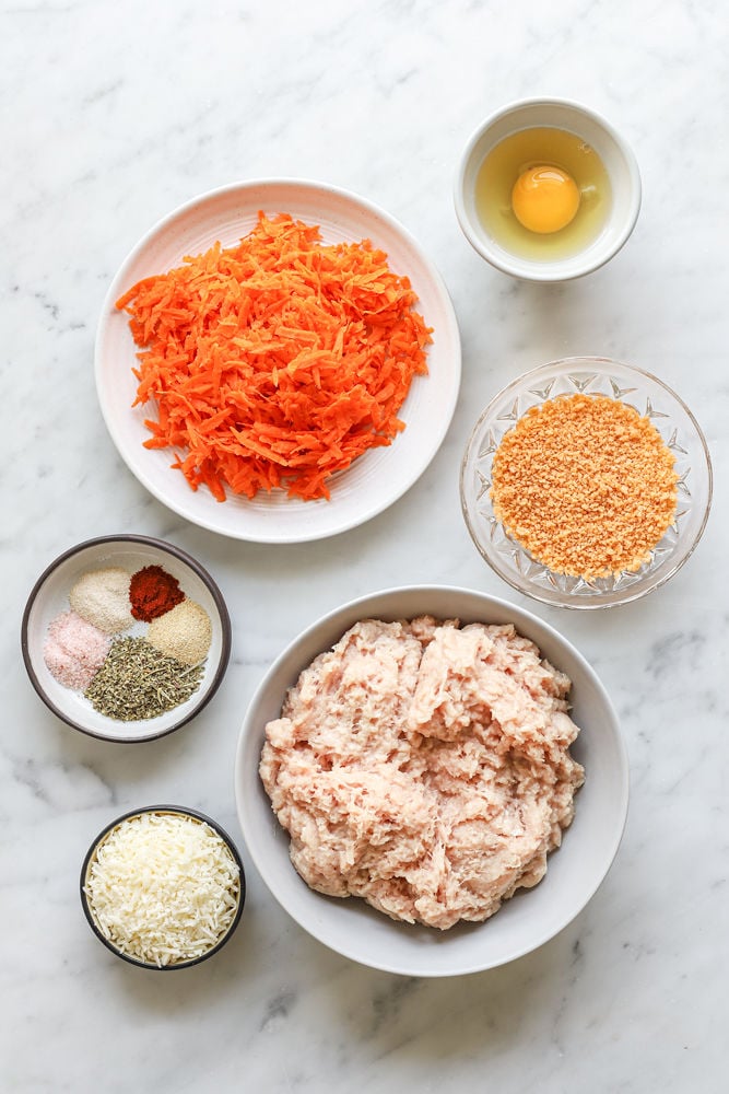 Ingredients for chicken carrot meatballs in bowls on a white table.