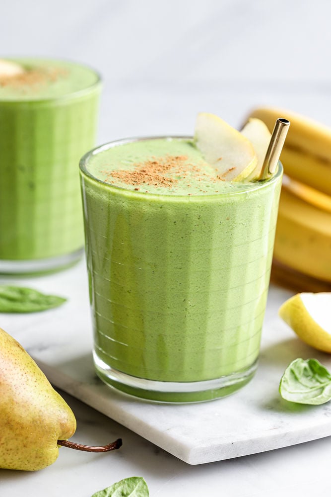 Spinach pear smoothie in a glass with a gold straw. There are sliced pears and cinnamon sprinkled on top. There is another smoothie blurred in the background and ingredients for the smoothie scattered around the glass. 