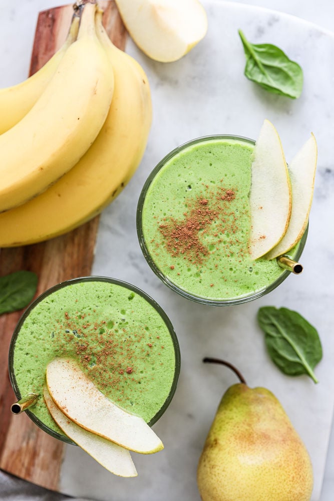 Two spinach pear smoothies in glasses, with metal straws and topped with sliced pear and cinnamon. There is banana, spinach, and pears next to the glasses.