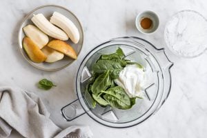 Spinach and yogurt and milk in a blender before blending. Pear and banana on a plate to side of the blender. A small bowl with cinnamon is also next to the blender.