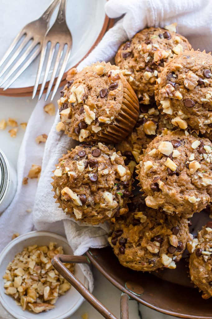 Banana zucchini muffins in a tray with a white napkin. There are plates with forks next to them and a small bowl with chopped walnuts.