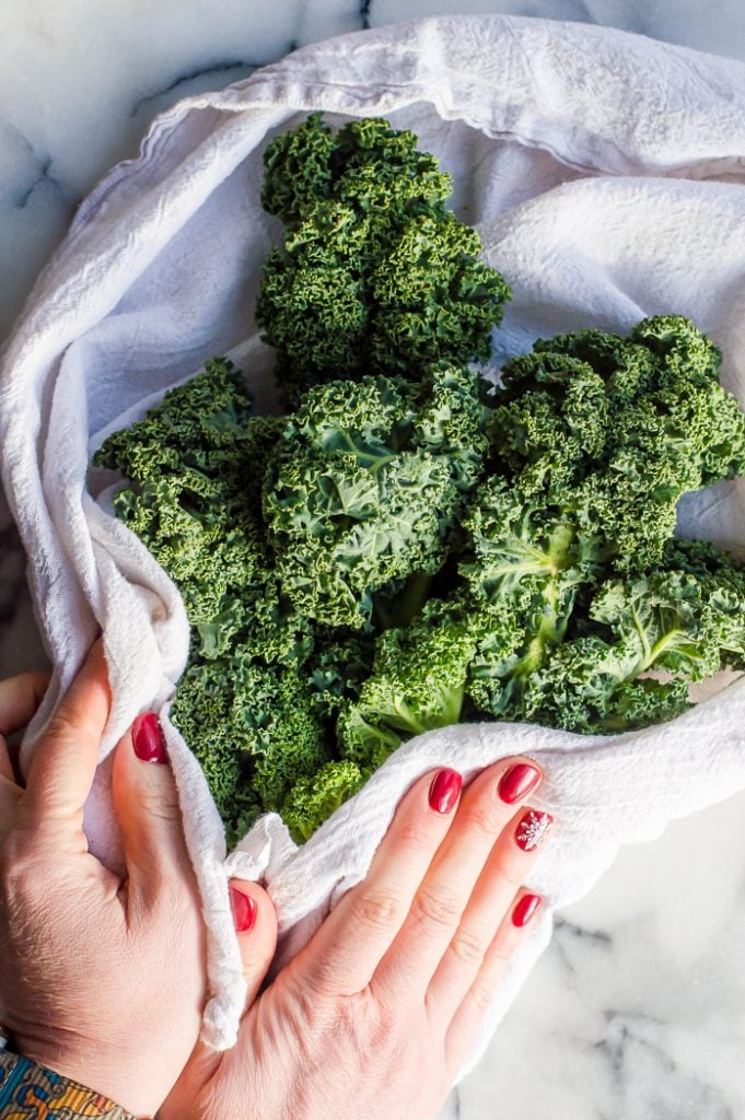 Two hands with red nail polish patting kale dry with a white tea towel