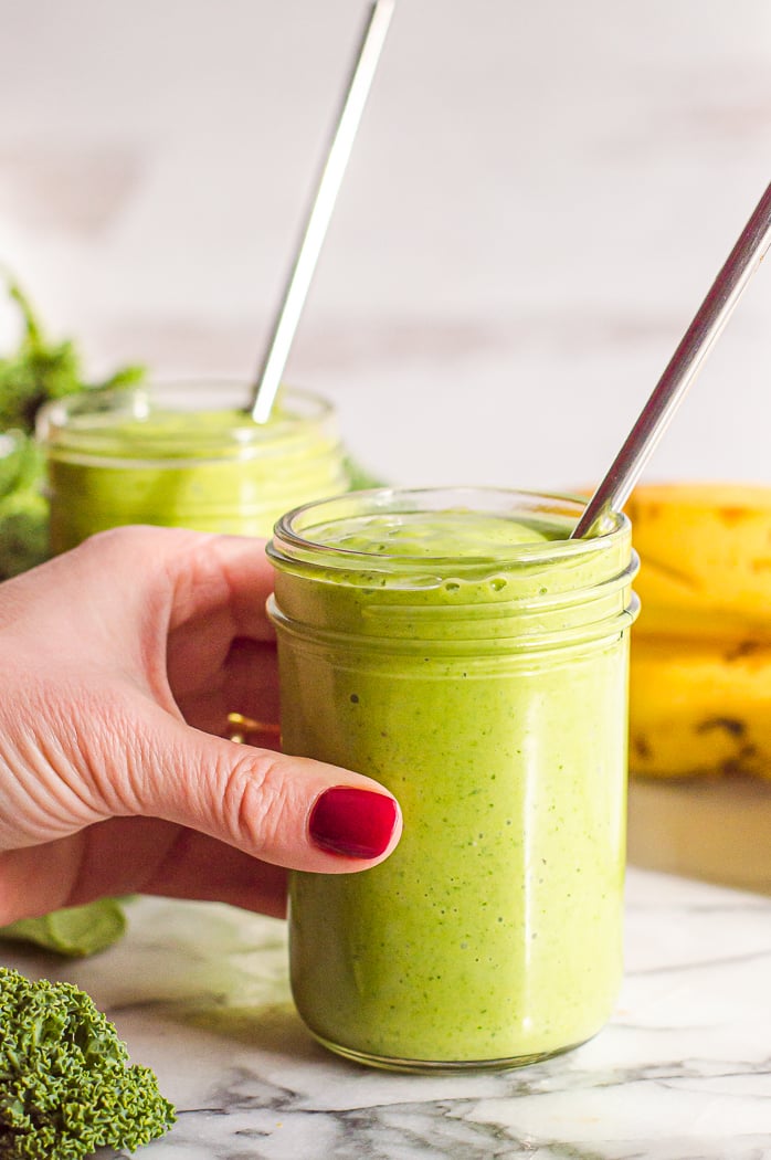 Kale and spinach smoothie in a jelly jar with a metal straw. A hand with painted red nails is reaching for the glass. There is another smoothie and ingredients blurred in the background. 