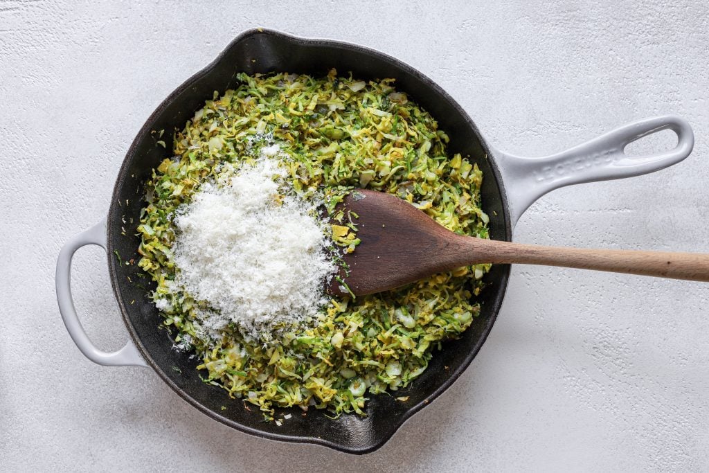 Le Creuset pan filled with finely chopped (shaved) Brussels sprouts and grated Parmesan cheese on top. Wooden spoon in the shaved Brussels sprouts (as if stirring).