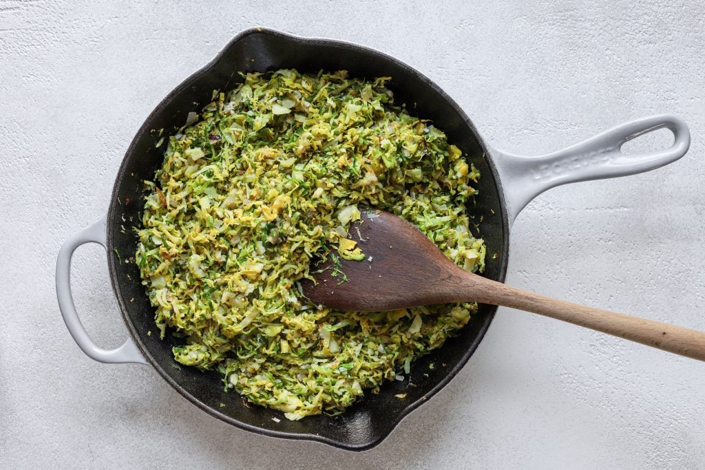 Le Creuset pan filled with finely chopped (shaved) Brussels sprouts mixed together with minced garlic and grated Parmesan cheese. Wooden spoon in the shaved Brussels sprouts mixture (as if stirring).