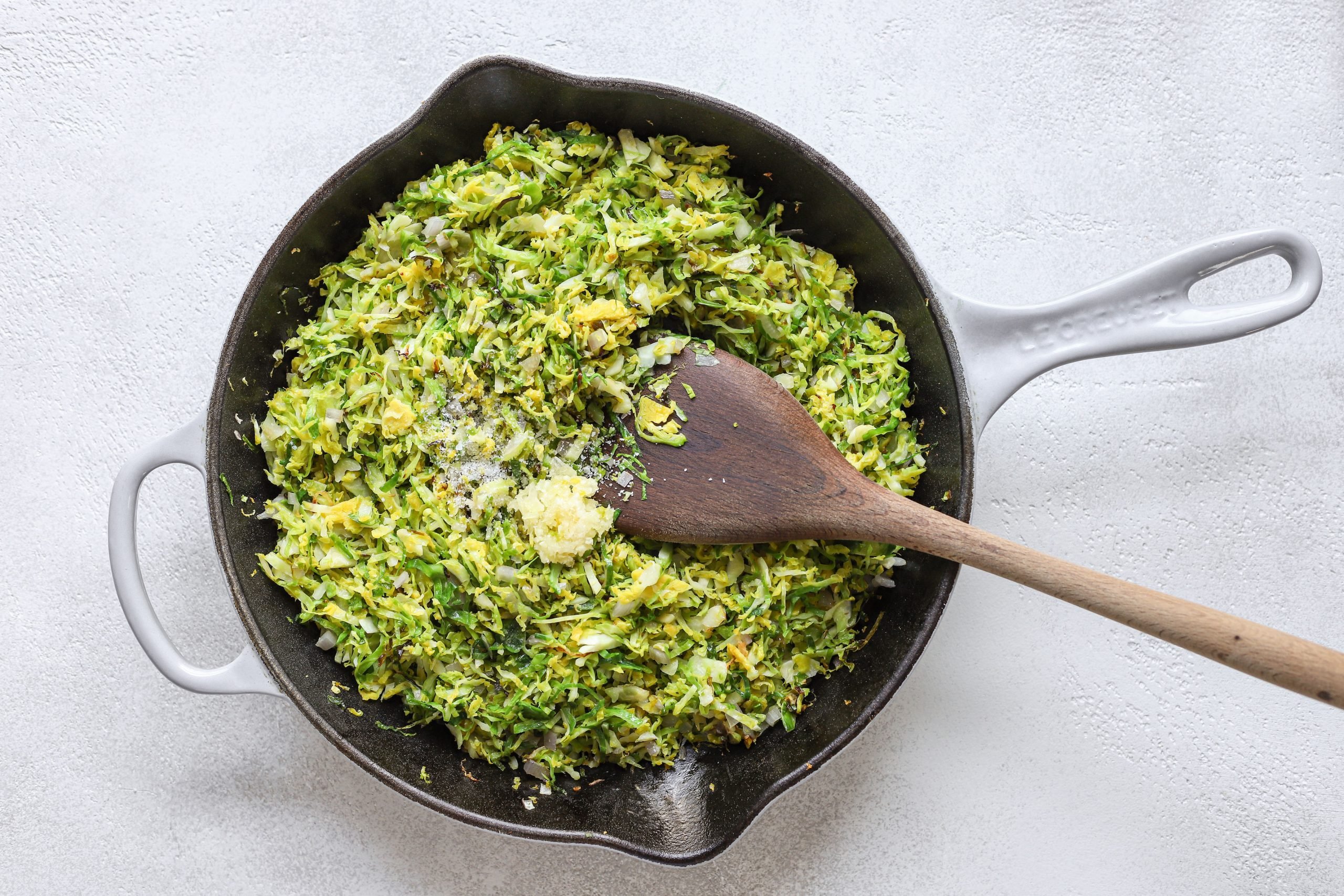 Le Creuset pan filled with finely chopped (shaved) Brussels sprouts and minced garlic on top. Wooden spoon in the shaved Brussels sprouts (as if stirring).