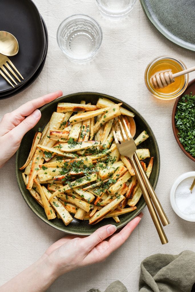 Honey roasted parsnips in a green bowl with gold serving utensils on a table. Two hands are holding the bowl. There is a bowl of honey with a dipper, a bowl of chopped parsley, and a bowl of salt on the table next tot he bowl of parsnips.