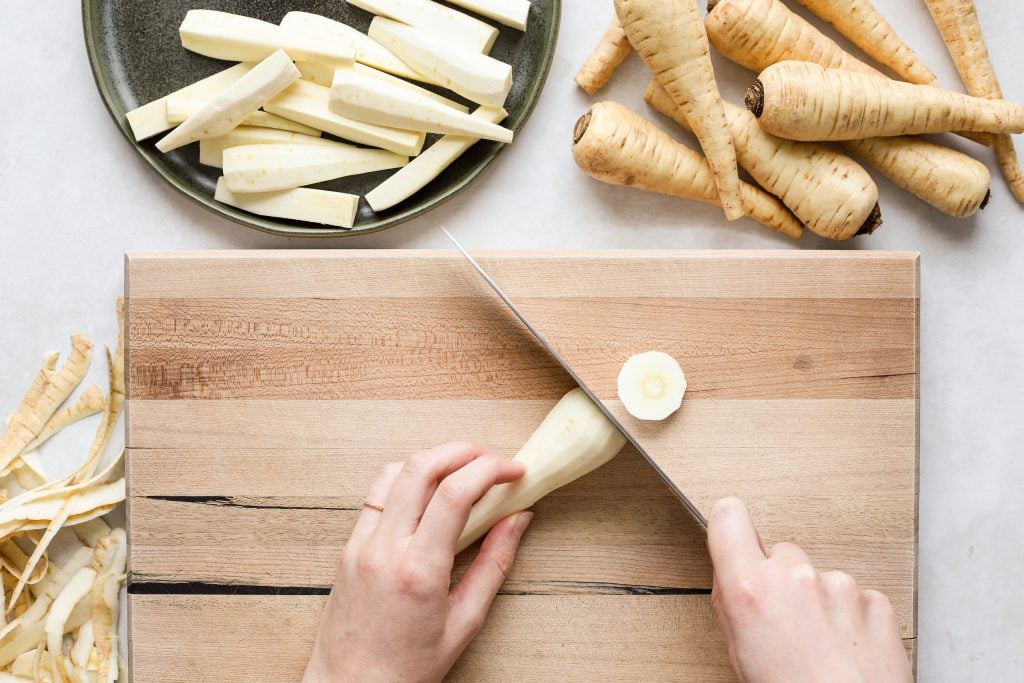 A hand trimming the top off of a peeled parsnip on a cutting board.