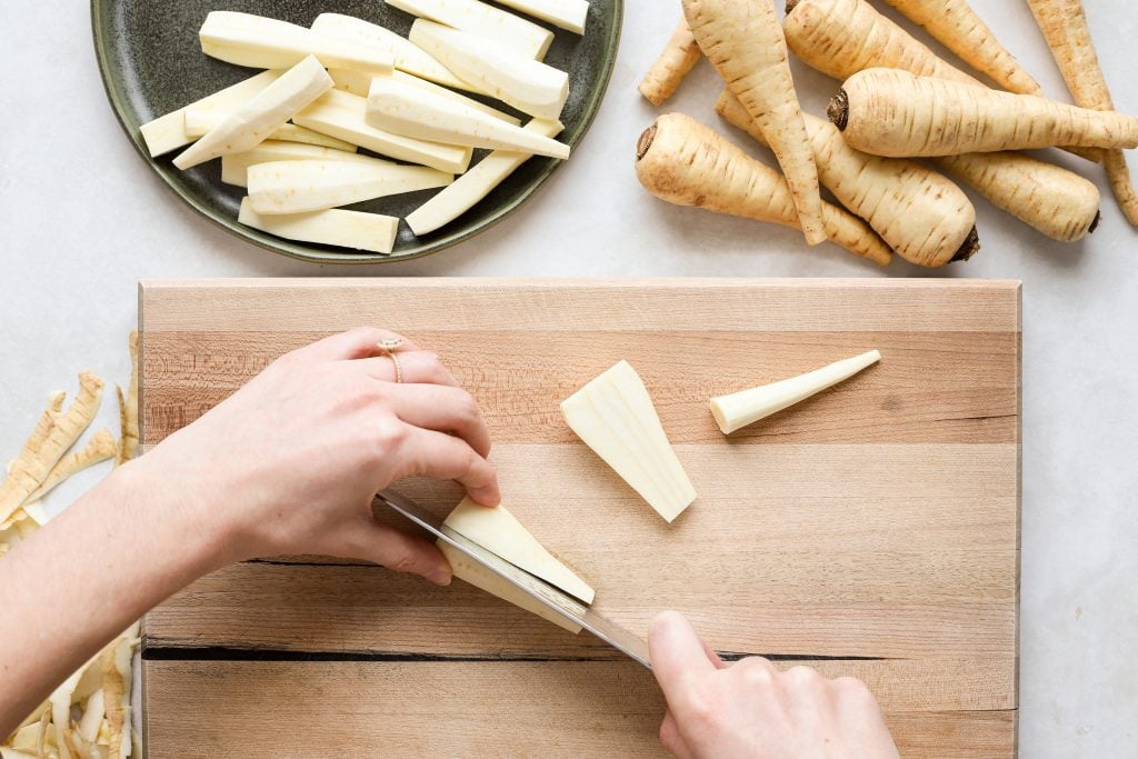 A person cutting a peeled parsnip into a fry shape on a cutting board.