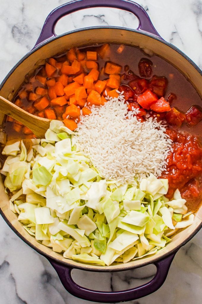 Chopped cabbage, uncooked rice, crushed and diced tomatoes, and diced carrots on top of the soup in a pot before being stirred in with a wooden spoon. 
