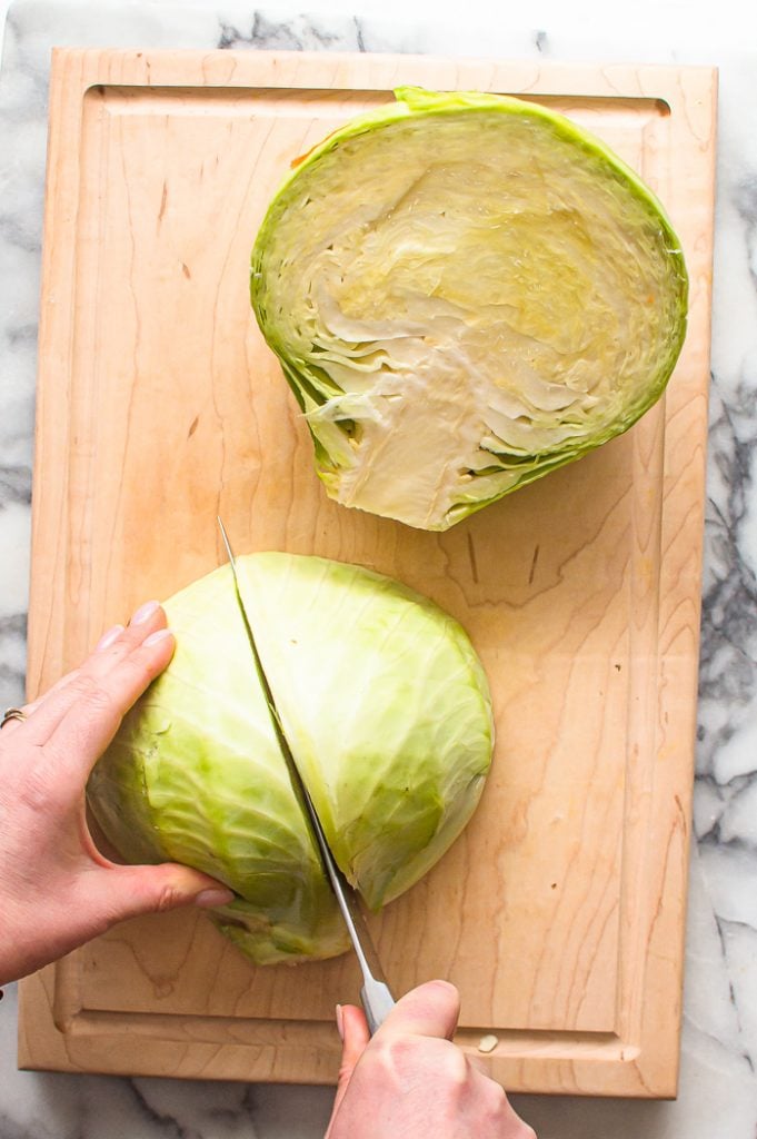 A hand holding a halved cabbage and another hand cutting the half into two pieces. 