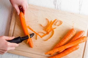 A person peeling a carrot over a wooden cutting board. There are other carrots on the cutting board.