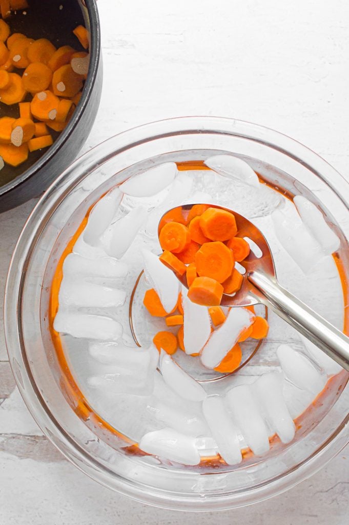 Slotted spoon pulling carrots sliced into coins out of ice water in a glass bowl. Glass bowl is sitting on a white distressed surface. Pot of water with coin-shaped carrot slices is set to the upper left side of the image.Slotted spoon pulling carrots sliced into coins out of ice water in a glass bowl. Glass bowl is sitting on a white distressed surface. Pot of water with coin-shaped carrot slices is set to the upper left side of the image.