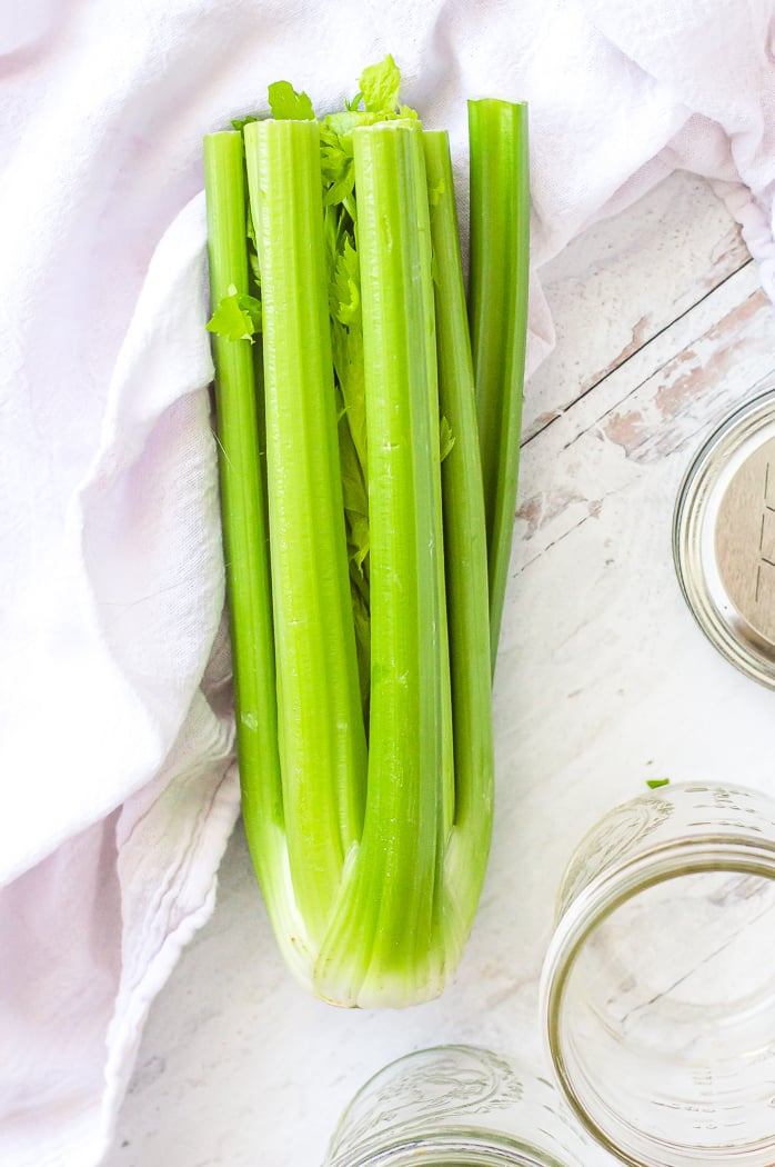 A whole bundle of celery is sitting on a white distressed countertop. Celery is in the center of the image. White tea towel is set to the side of the image on the left. Glass jar and metal lid are on the counter to the right of the celery.