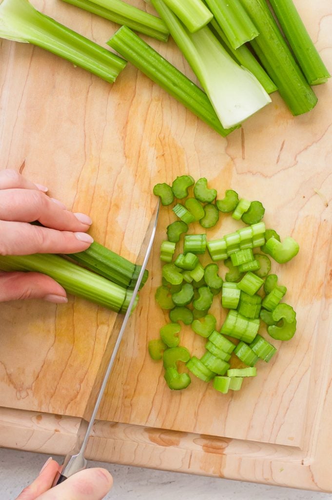 Hand chopping celery on a wooden cutting board using a chef's knife. Chopped pieces of cutting board are in a pile on the right side of the cutting board. Larger celery pieces are set to the side on the top of the cutting board.