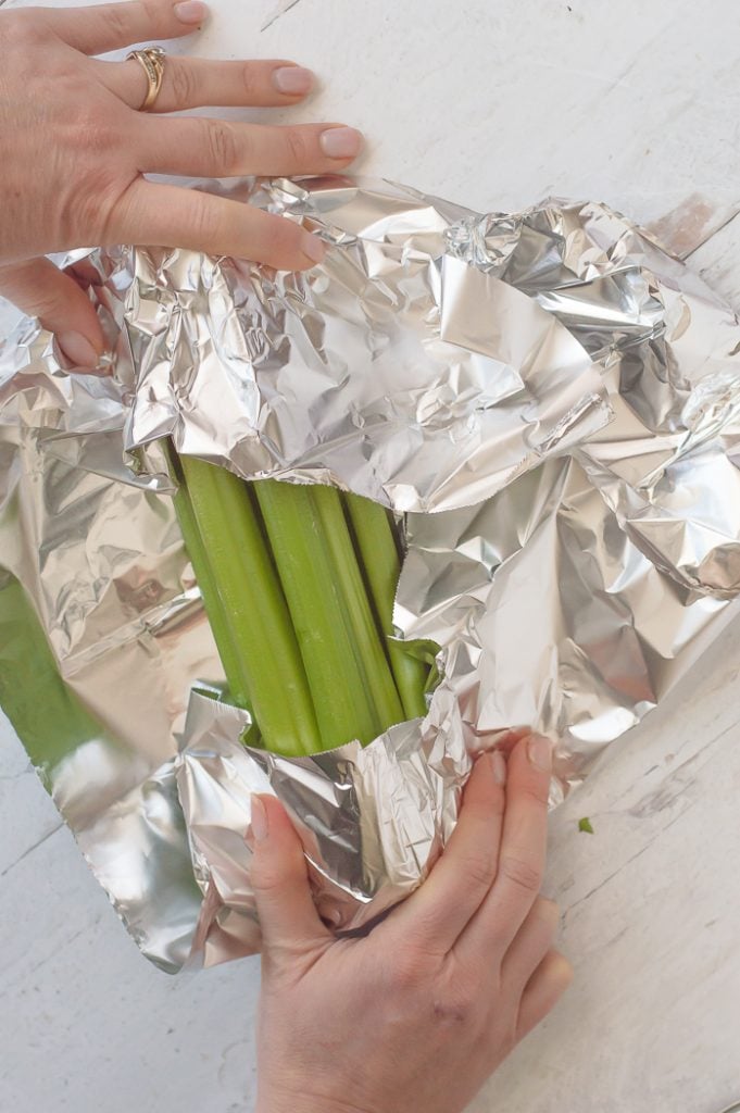 Two hands wrapping a bundle of celery in aluminum foil. Foil-wrapped celery is on a white distressed countertop.