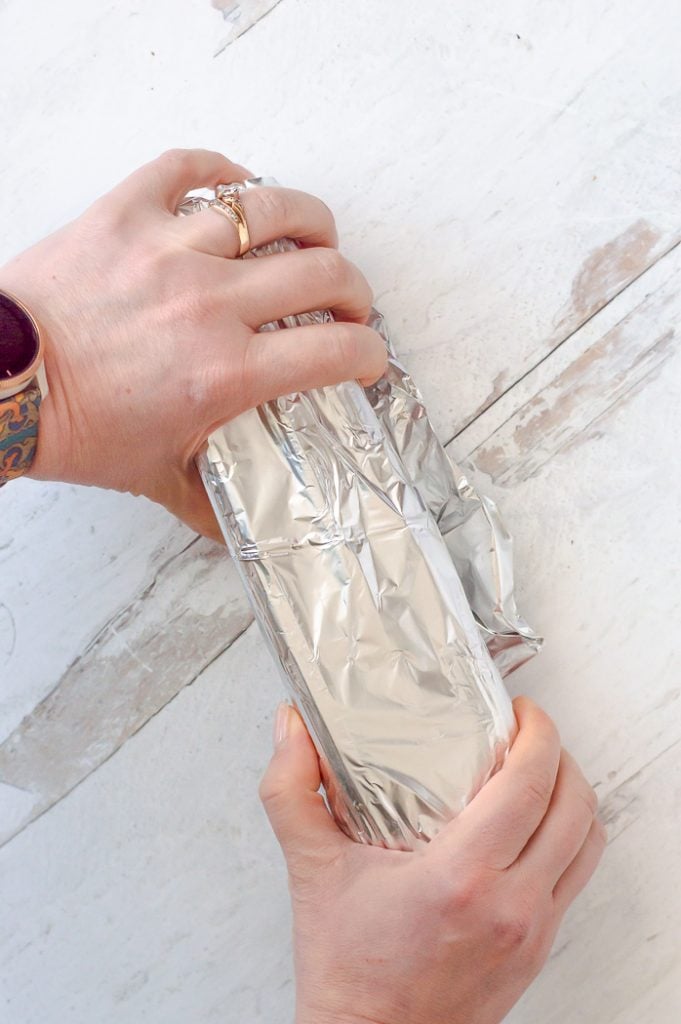 Two hands holding onto either end of a bundle of celery (celery is not visible) wrapped in aluminum foil. Foil-wrapped celery is on a white distressed countertop.