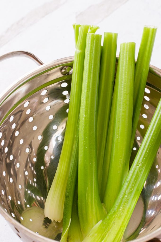 Six long stalks of celery in a metal colander. Colander is sitting on a white marble countertop.