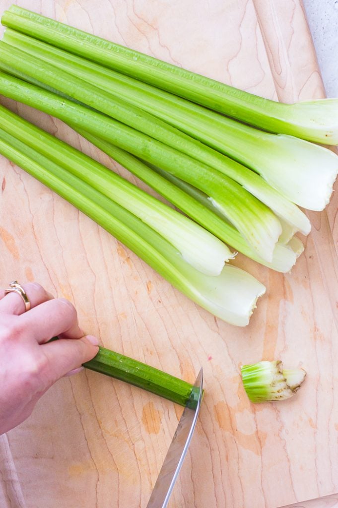 Hand holding one long stalks of celery while end is being trimmed off by a chef's knife. Trimmed end is set to the side on the right. Celery pieces not yet trimmed are at the top of the image. Celery pieces are being cut on a wooden cutting board.
