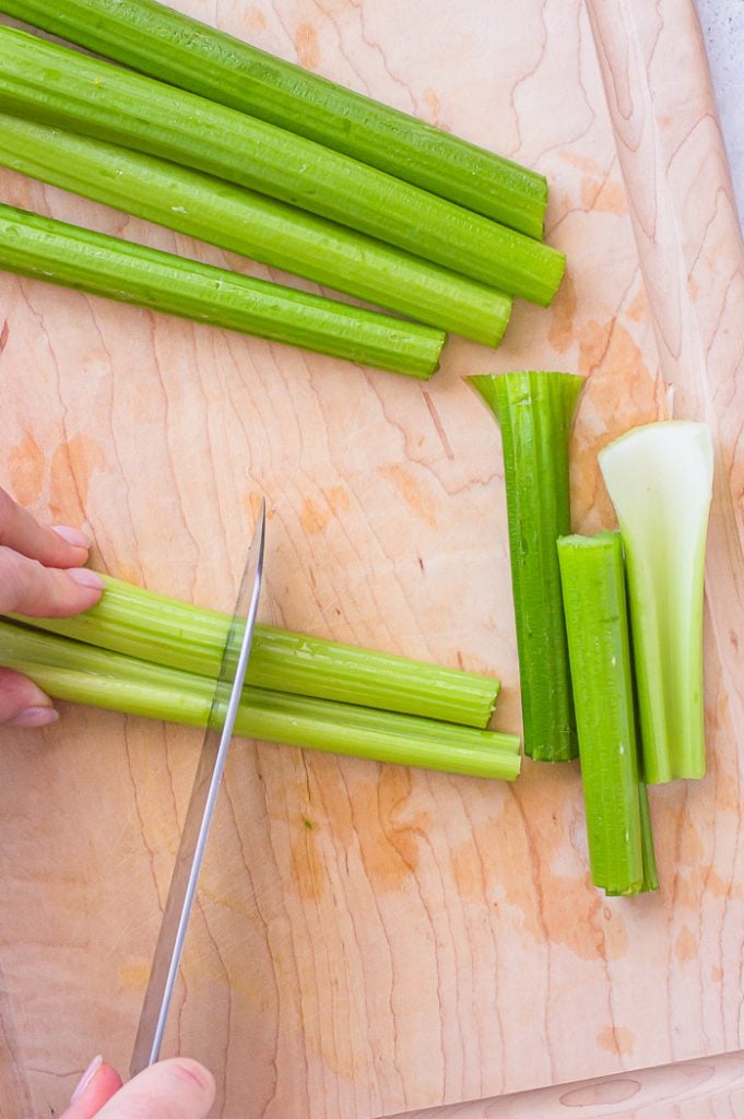Hand holding onto two long stalks of celery while being cut by a chef's knife. Cut celery pieces are set to the side on the right. Celery pieces not yet cut are at the top of the image. Celery pieces are being cut on a wooden cutting board.