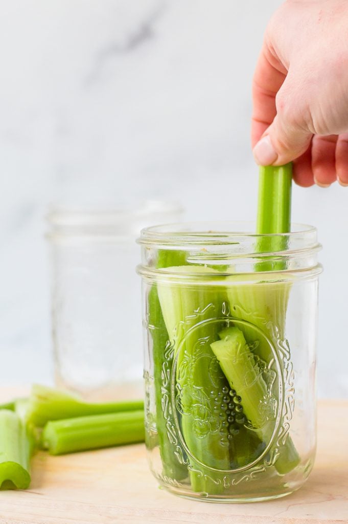 Hand pulling out one piece of trimmed celery from a bunch of pieces in a glass jar filled with water. Glass jar is sitting on a wooden cutting board. Additional celery pieces are set to the side on a cutting board. Additional glass jar is in the back of the image. 