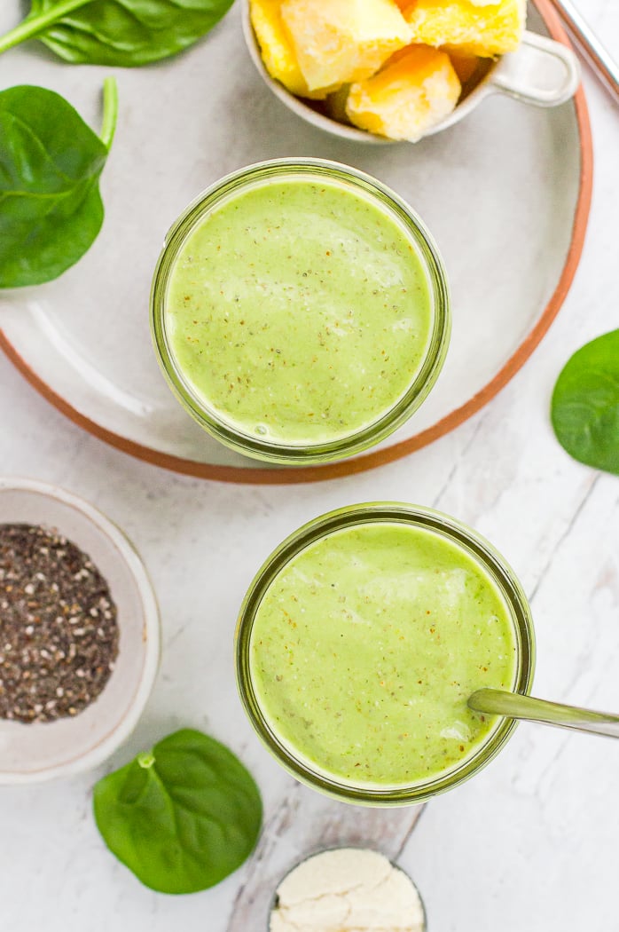 Green protein smoothie in two glasses. There are ingredients for the smoothie scattered around the glasses.