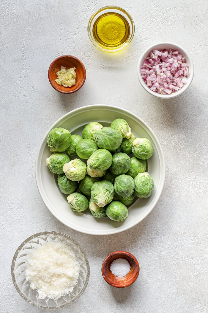 6 bowls of varying styles and sizes (glass, wood, ceramic) holding individual ingredients set on a white table. Small glass bowl container contains oil. Small wooden bowl contains minced garlic. Medium white bowl contains chopped red onion. White dish in the middle of the image with whole Brussels sprouts piled on top. Medium glass bowl with grated cheese. Small wooden bowl with salt.