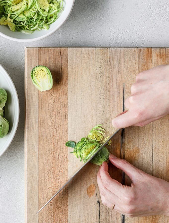 Two hands holding a chef's knife finely chopping half of a Brussels sprout on a cutting board. The remaining half of the Brussels sprout is set to the side on the cutting board. Two white bowls set to the side of the image. One bowl has whole Brussels sprouts. The other bowl contains finely chopped Brussels sprouts. 